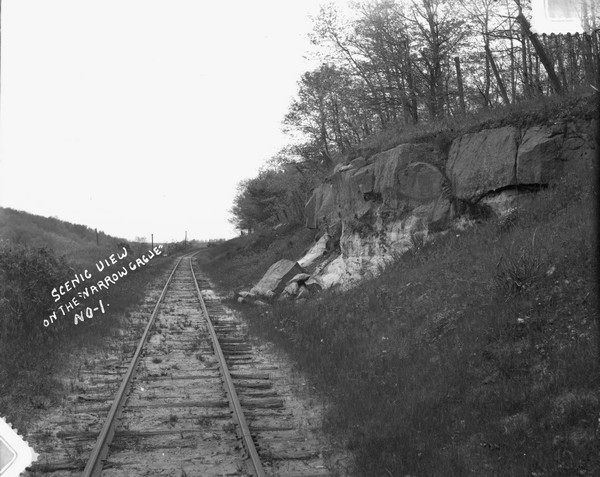 View down middle of narrow gauge railroad track. On the right is exposed rock on side of tree-lined hill.