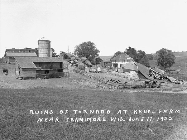 View down hill of the aftermath of a tornado at a farm. There are two collapsed barns, and an intact silo, outbuildings and farmhouse. A horse is standing on the far left.