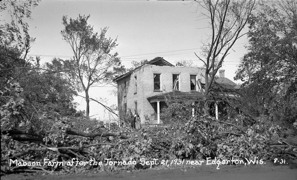View across felled trees towards two women standing near the porch of a damaged brick house.