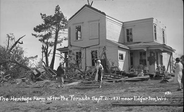 View of damaged farmhouse and group of people sitting and standing outdoors. The roof was blown off and windows were shattered. Four women stand in the yard near fallen trees, a hand-pump and debris. Gathered on the porch are two women, one holding an infant, two men, one with a dog, and a young child.