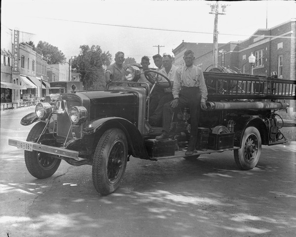 Five men posed on a firetruck in the middle of a street in a business district. Storefronts are in the background on the left and right.