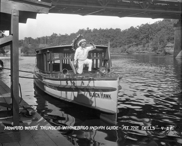 View from pier of a Native American man wearing a headdress kneeling on a tourist boat called "Black Hawk" docked under a bridge in the Wisconsin River. He is holding a megaphone that says "Black Hawk." Trees and rock formations are on the opposite bank.