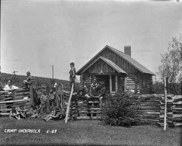 A group of young boys with their male camp counselors are sitting and standing around the split-rail fence. There is a log cabin and a cornfield in the background.