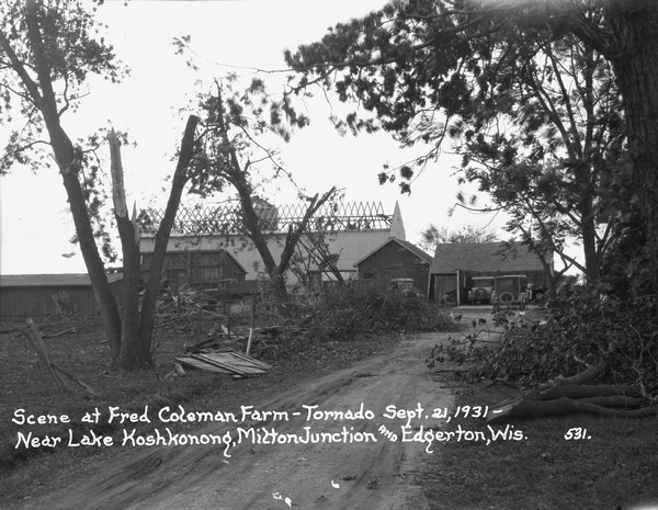 View from road of storm damage to a farm. The barn roof has blown off. Men are working on the roof, which has exposed roof timbers. In the foreground are trees with broken limbs. Pigs and chickens are in the barnyard. There are a number of parked cars and trucks near outbuildings. Another building on the far right may be a farmhouse.