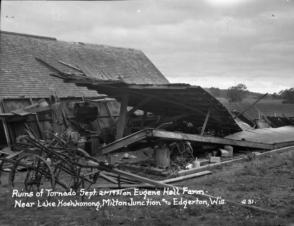 Destroyed farm building next to a standing barn. Agricultural equipment is in the foreground.