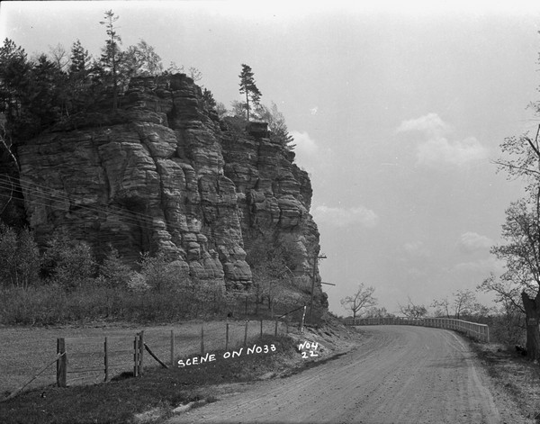 View from road of a bluff with exposed rock face next to a winding unpaved road (probably Hwy 33 up to Wildcat Mountain). There is a guardrail along the curve in the road on the right. There is wire fencing along the road on the left.