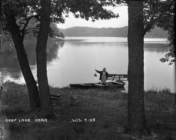 View down slope towards Deep Lake. There is a girl standing in a rowboat at the shoreline. She is holding up her catch of the day. In the background a man is wading along the shoreline on the left.