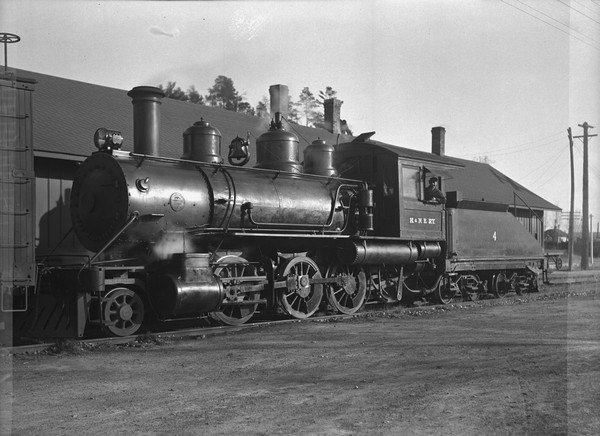 Locomotive at the depot with what may be a coal car. The engineer is leaning out the window.