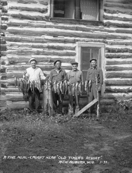 Group portrait of four men standing behind a row of fish on a stringer. Two large muskies are in the center. The men are standing in front of a two-story log cabin.