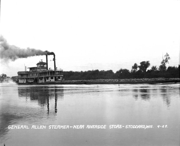 View across water of the <i>General Allen</i> steamer pushing barges down the Mississippi River. Men are on the steamer, and two more men are standing on the barges which are loaded with unknown material.