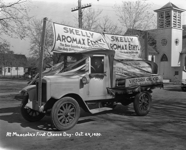 Man sitting in driver's seat of a Skelly Truck promoting itself with banners and streamers. In the background on the right is a church.
