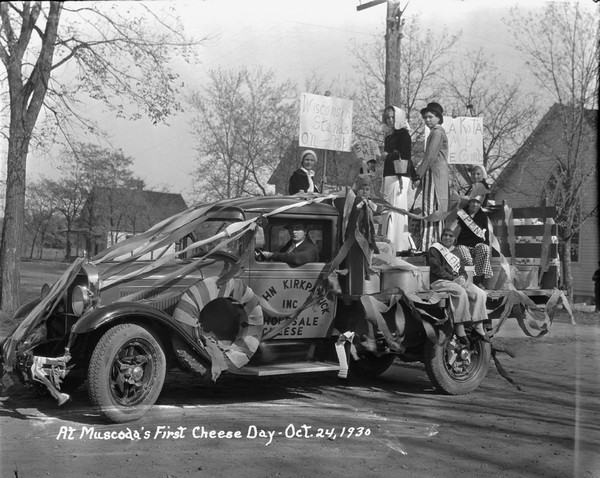 Group portrait of children in costume posed on the back of a truck decorated with streamers for the Cheese Day Parade. Some of the children hold handwritten signs. A man sits in the driver's seat. There is a church building in the background on the right. A sign on the driver's side door reads: "John Kirkpatrick Inc., Wholesale Cheese."