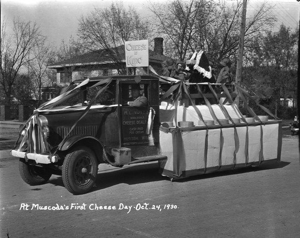 Group portrait of children posed on flatbed truck decorated with streamers as a float for Cheese Day. A sign reads: "Cheese is King," and a boy dressed as a king wearing a crown and robe, is surrounded by children in costume. The sign on the driver's side door reads: H.L. Noyes, Wholesale Cheese Dealer."