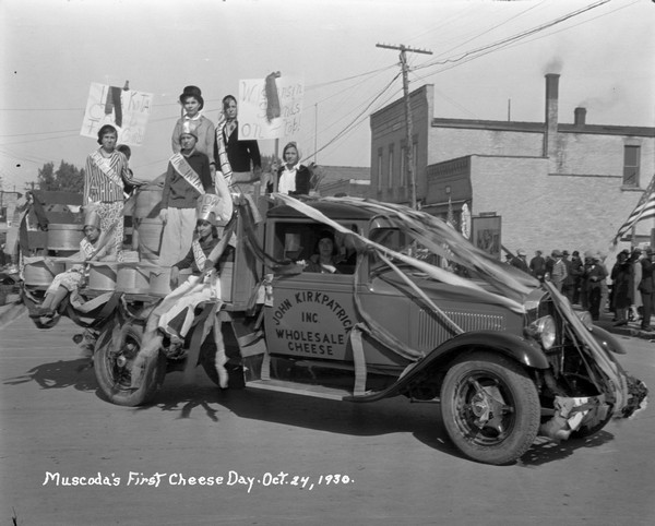 Group of people posing among round wood cheese boxes on a flatbed truck decorated as a float in the Cheese Day Parade. One person holds a sign that says: "Lakota Camp for Girls," and another sign says: "Wisconsin Stands on Top." One girl wears a sash that says: "Wisconsin" and other girls wear sashes for other states. The sign passenger side door reads: "John Kirkpatrick Inc., Wholesale Cheese." Spectators are watching from the sidewalk on the right.