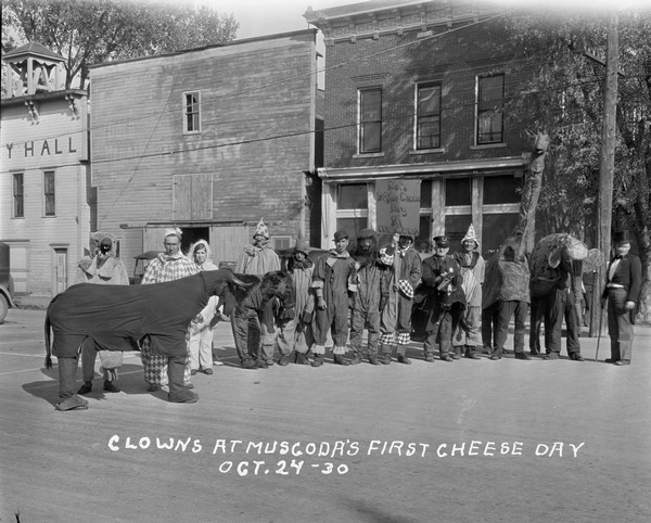 A line of clowns and assorted pantomime animals dressed up for Cheese Day. The animals include; a horse, a woodchuck, a bear, a giraffe and an elephant. A man dresses as a police officer is standing in the center, and a man next to him is holding a sign that says: Day After-Cheese Day at Muscoda." The buildings behind them are a bakery, beauty shop, livery, and on the far left, City Hall.