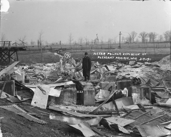 A man stands on a foundation among the debris of the aftermath of a powder explosion. Around the man are pieces of corrugated metal, machinery and wood. In the background an elevated pipe or fence runs across a field.