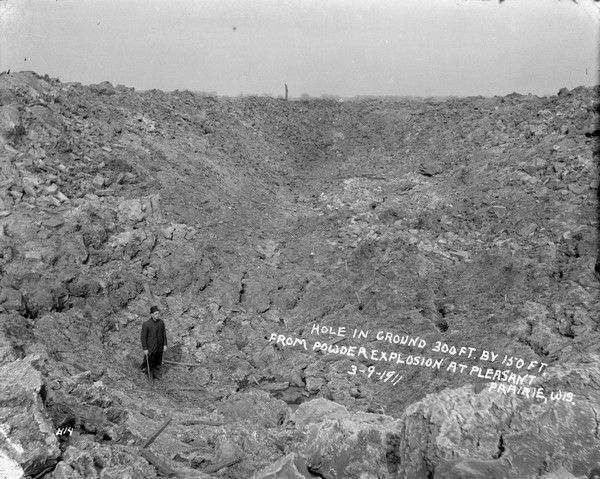 View looking down of a man standing among rubble and other debris at the bottom of a "300 ft. by 150 ft." hole in the ground from a powder explosion.