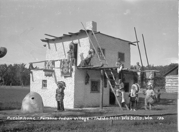 Eight Native American men pose around the two-story stucco sided Pueblo Home. They are all wearing traditional dress, with four wearing headdresses, and one man holding a drum. Two rugs are hanging over the balcony rail, and another hangs over a timber near the entrance. A ladder leans against the facade for access to the balcony. On the far left is an arched-shaped structure that may be an outdoor cooking fireplace. A child stands near a log cabin in the background on the right.