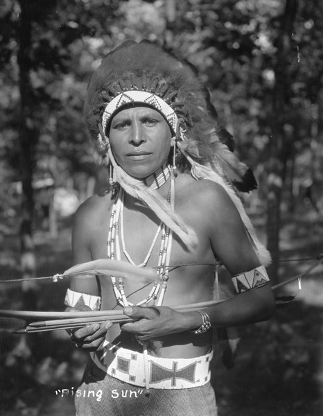 Waist-up outdoor portrait of a Native American man wearing strings of beads, a silver bracelet and a headdress. He is holding a bow and arrows.