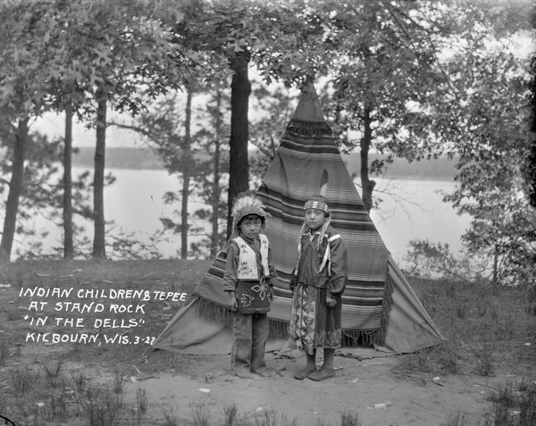 A Native American boy and girl pose standing in front of a tipi on a hill by a lake. The tipi is covered with a fringed blanket.
