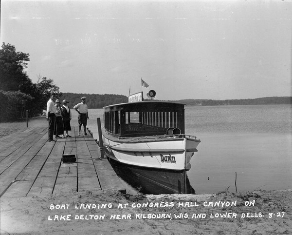 Four people, two men and two women, stand on a pier near an excursion boat on Lake Delton. A sign with flags is on top of the boat and reads: "Lake and River Front Lot," and the name of the boat is <i>Dawn</i>.