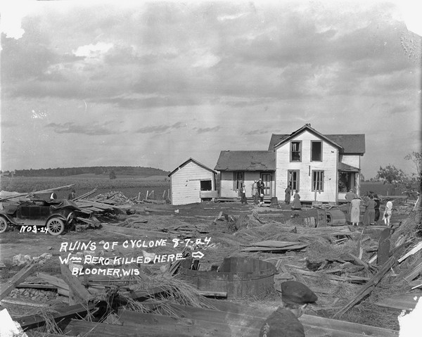 The aftermath of a killer tornado, including an arrow and caption that reads: "William Berg Killed Here." In the background near the farmhouse are a group of people and chickens. The farmhouse windows are blown out and shingles have been ripped from the roof. Broken pieces of wood and other debris are strewn around the yard. There is a young child in the foreground and two men stand looking at debris behind a damaged automobile parked on the far left.