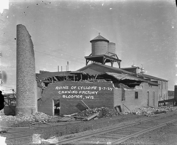 View across railroad tracks of the canning factory in the aftermath of a tornado. The top of the brick structure and the smokestack has been blown off. Piles of bricks lay at the base and cover part of the railroad tracks. The water towers and a wood building behind have no visible storm damage.