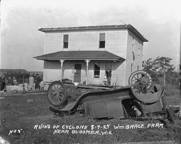 View across yard of a storm-damaged farmhouse. In the foreground is an overturned automobile. The windows of the house have been blown out and the roof has completely blown off. A boy and girl stand on the porch, and a group of people standing around a broken hand-pump on the left.