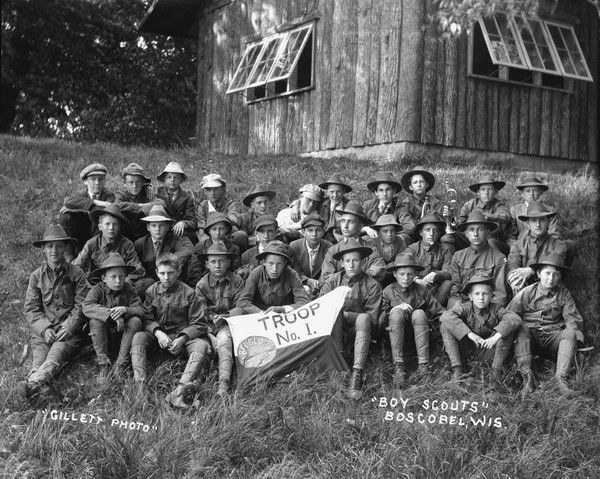 Group portrait of Boy Scout Troop No. 1 of Boscobel. Three rows of boys and their scout masters are sitting on a hill in the tall grass in front of a log building. A boy in the front row holds the banner. There are two buglers in the back row.