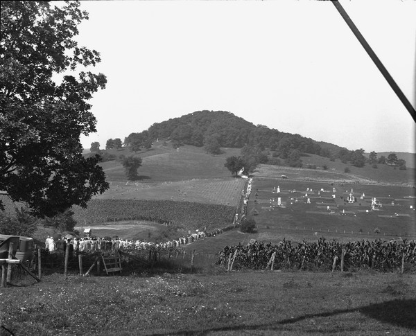 View down valley of a procession of hundreds of pilgrims walking up the hill to St. Anne's Chapel which is lined with Stations of the Cross. The crowd is walking on a path among fields and a cemetery. Automobiles are parked near a fence on the left.