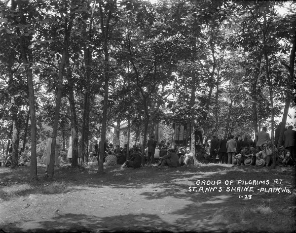 Pilgrims gathered outside the shrine. Many of the people are sitting on the ground among the tree. There are banners hanging in front of the stone chapel.