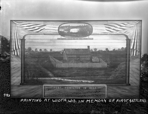 Painting of Fort Hamilton propped outdoors on a woodpile. An oval inset painting at the top commemorates is titled: "Col. Hamilton Leaving For The Gold Fields 1849." A sign at the bottom is titled: "Fort Hamilton in 1832." It reads: "Built by Colonel William Stephen Hamilton, youngest son of the renowned American Statesman, Alexander Hamilton. Col. Hamilton was the founder of Hamilton's Diggings - now Wiota. He was born in New York Aug 4, 1797. He spent three years at West Point after which he was appointed on the staff of William Rector, coming west to Illinois, and landing at Galena on July 4, 1827. In 1828 he started lead mining and smelting at Hamilton's Diggings. Col. Hamilton was a prominent citizen of this territory and a Captain in the Black Hawk War. He left for California in May 1849 after the gold discovery, and died at Sacramento in 1852."