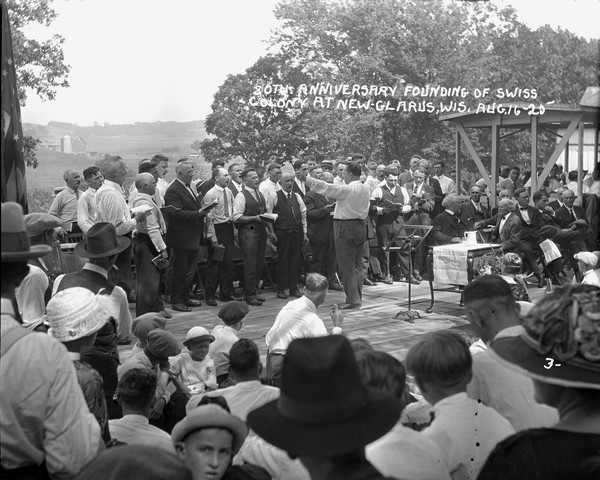A chorus of men singing on stage with a crowd watching. A group of officials seated on the right. Rolling farmland in the background.
