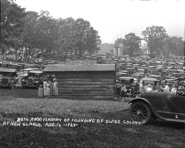 View of field full of parked automobiles at the commemoration of the 80th anniversary of the founding of the Swiss Colony at New Glarus. A log structure in the foreground has a sign on top that reads: "New Glarus Settled 1845." People are picnicking under the trees. A farm with two silos is in the background.