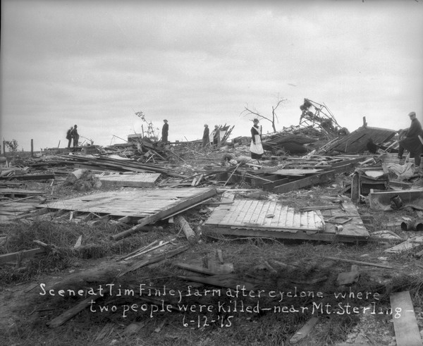 People standing among the broken remains of the Tim Finley farm after a killer tornado. A woman in the center is standing on a mattress. Two people were killed.