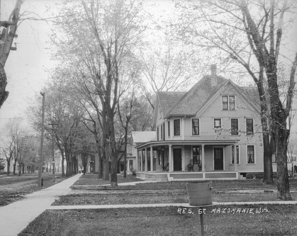 View down row of residential houses along a sidewalk. The first house has a wrap-around open porch with columns on the front and side. In the distance two women are walking up the block on the sidewalk.