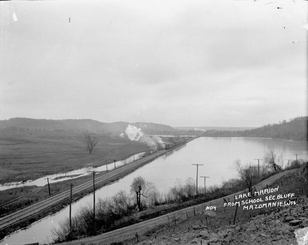 Elevated view of Lake Marion, and the railroad tracks that run along it on the left. A train belching smoke is coming up the tracks. A road runs along the lake in the foreground.