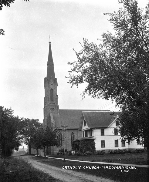 View across unpaved road of the Catholic Church, which is a stone building. Includes a tall steeple a cross on top. Narrow, pointed arched windows are on the side. A parsonage with a vine-covered porch is on the right.
