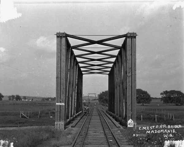 View down railroad tracks of a railroad bridge, which has framed beams on the sides and top. A boy and girl are standing and posing on the bridge. Signs are handwritten at the base and top and read, in part: "No. 1n, No. 2n, No. 3n, No. 4n." Another sign says: "B 400." Farms, fields and a bluff are in the far background.