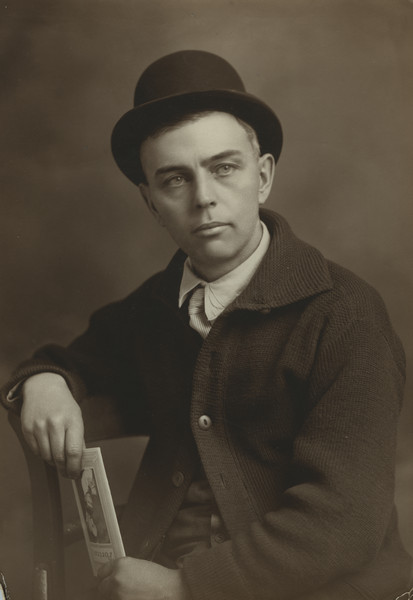 Waist-up portrait of Sherwin Gillett sitting in a chair, wearing a sweater, striped necktie and a hat. He is holding a brochure titled: "Portraits."