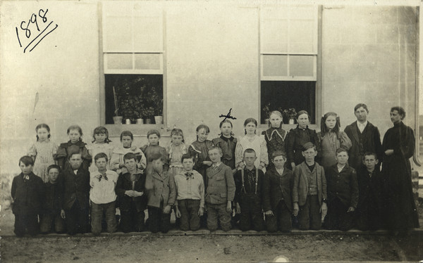 Outdoor class group portrait of the 5th and 6th grades of Muscoda Public School. An "X" marks Emma Schmitt (the future Mrs. Sherwin Gillett). Boys kneel in a row in front, the girls stand in a line behind. The teacher, a woman, stands on the far right, next to an older boy or man standing to her right. Potted plants are lined up along the windowsills of the open windows behind the group.