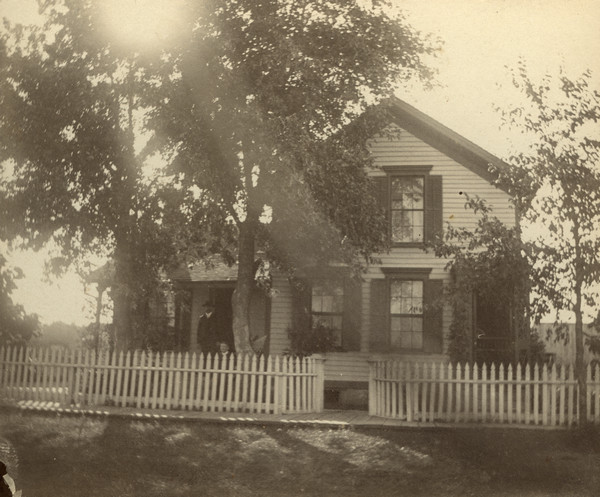 View of the exterior of the Conrad Schmitt (Sherwin Gillett's future father-in-law) residence. There are two unidentified men on the porch posing in the shade of trees. A wooden sidewalk and picket fence are in front of the house.