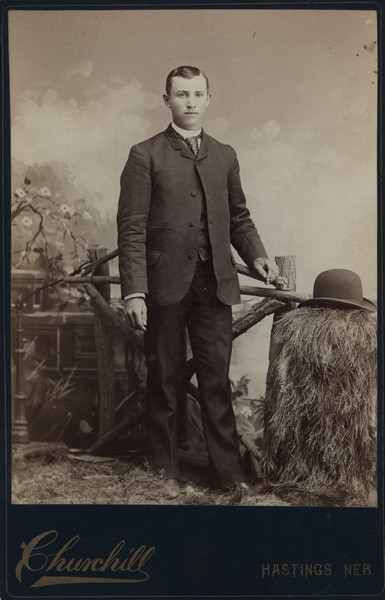 Full-length portrait in front of a painted backdrop of Dr. Charles Schmidt (Sherwin Gillett's wife's cousin). He is standing next to a prop fence and his hat rests on what appears to be a pile of hay. In 1910 he was a veterinarian living in Dodgeville.