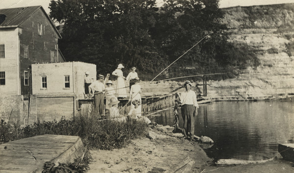 Photographic postcard along shoreline of a group of people fishing at Rudolph Mill. Sherwin Gillett's wife's cousin, William Schmitt, is standing in the foreground with his catch of the day.