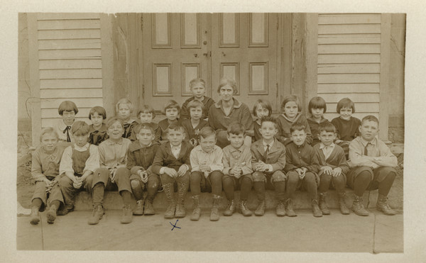 Outdoor group portrait of a class featuring Lorin Gillett (son of Sherwin Gilett), and their teacher sitting on the steps in front of the double door of what is probably the school building. Lorin would be about 8-years-old here.