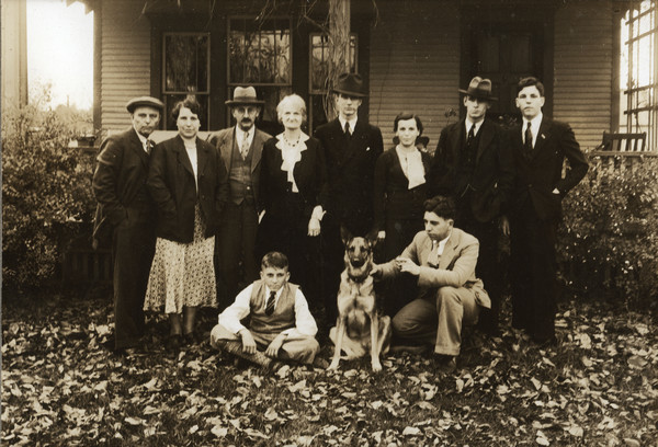 Group portrait on the lawn in front of a porch at the 25th Wedding Anniversary of Sherwin and Emma Gillett. Sherwin and Emma are standing on the left. Their sons, Marion and Harry, are posing on the ground in front with the dog. Daughter Cherye is standing behind Harry. Son, Lorin is standing on the right. Four other people are unidentified.