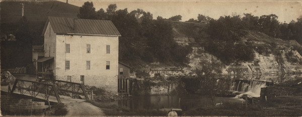 View of the Rundolph Mill and dam near Muscoda. A man stands near the far side of the bridge on the left. The banks of the river are rocky cliffs, and a steep hill rises in the background on the left.