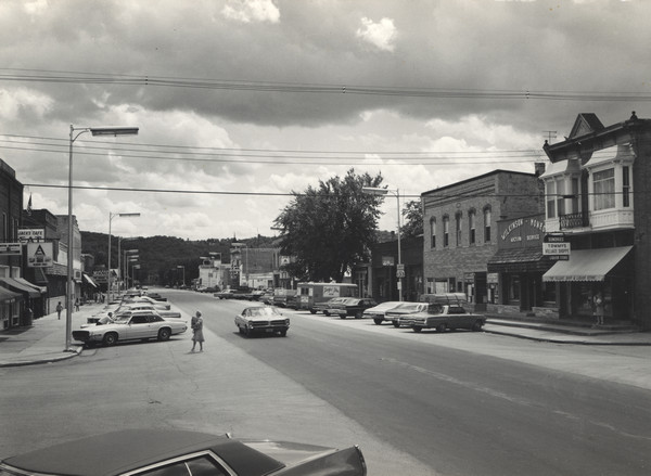 Slightly elevated view of a central business district. The theater marquee on the left reads: "The Concert for Bangladesh." Across the street on the right are storefronts, including Millers Market, Wilkinson-Monroe Auction Service, and Tommys Village Shop. Further down the street is a Skelly service station. Automobiles are parked at an angle along the curbs, and pedestrians are along the sidewalk and street. The tree-lined ridge of a bluff is in the far background.