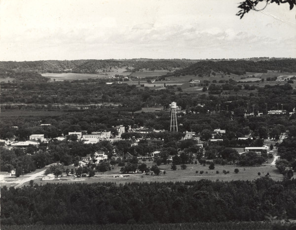 Elevated view over trees of the town of Muscoda. The water tower is in the center. The commercial district is on the left, and dwellings are in the foreground. Fields, the Wisconsin River and bluffs are in the far background.