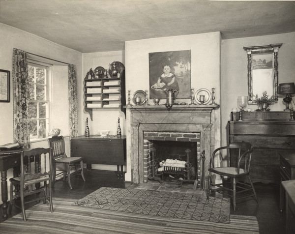 An interior view of a room in Pendarvis House. To the right of the fireplace is a dresser in the corner. On the left of the fireplace is a drop-leaf table. Another table is against the left wall near a window. Above the fireplace is a painting of a girl with a cat.